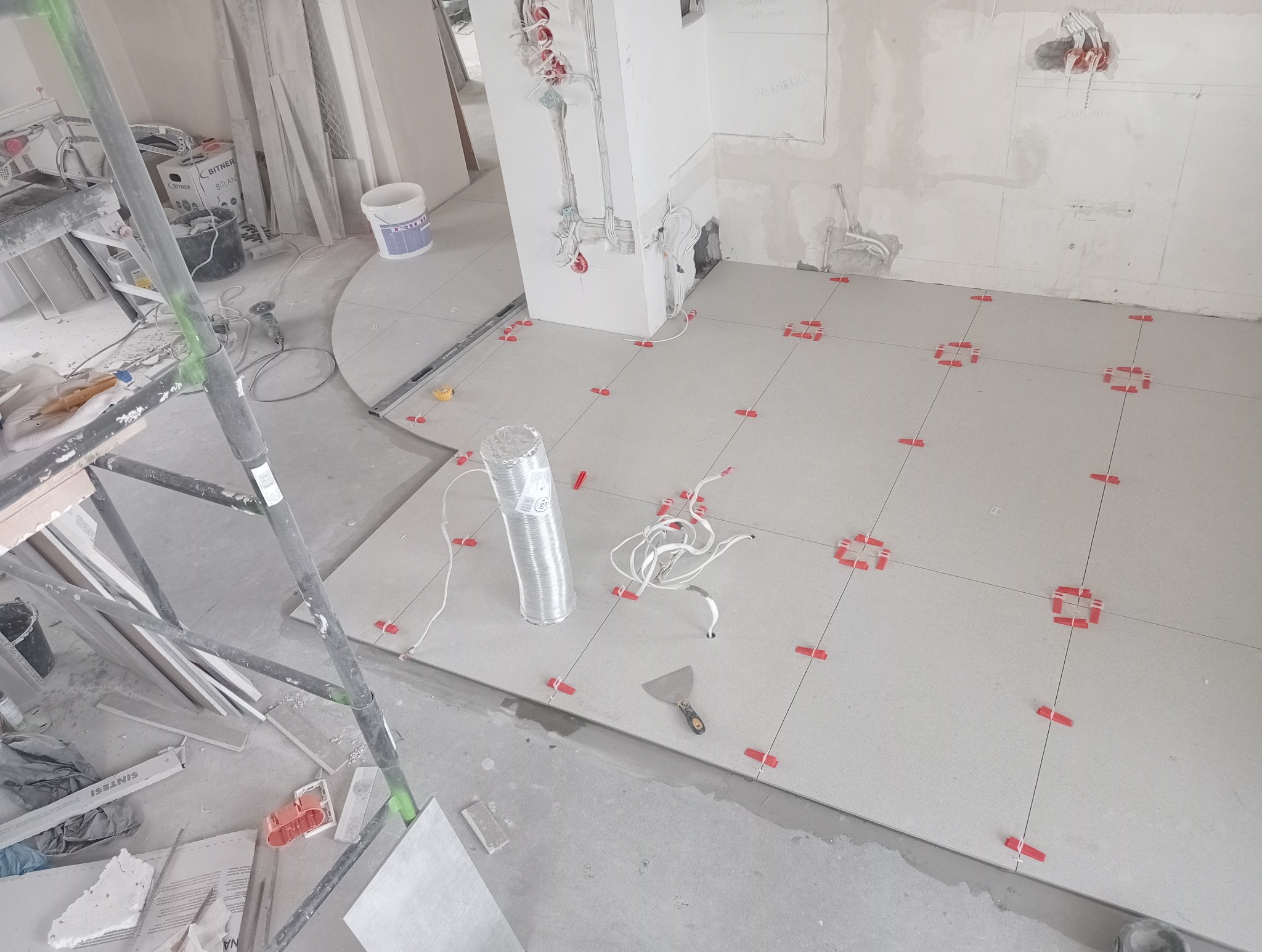 Jasne płytki podłogowe w trakcie układania, z czerwonymi krzyżykami dystansowymi, widoczne narzędzia i instalacje elektryczne w surowym wnętrzu.
