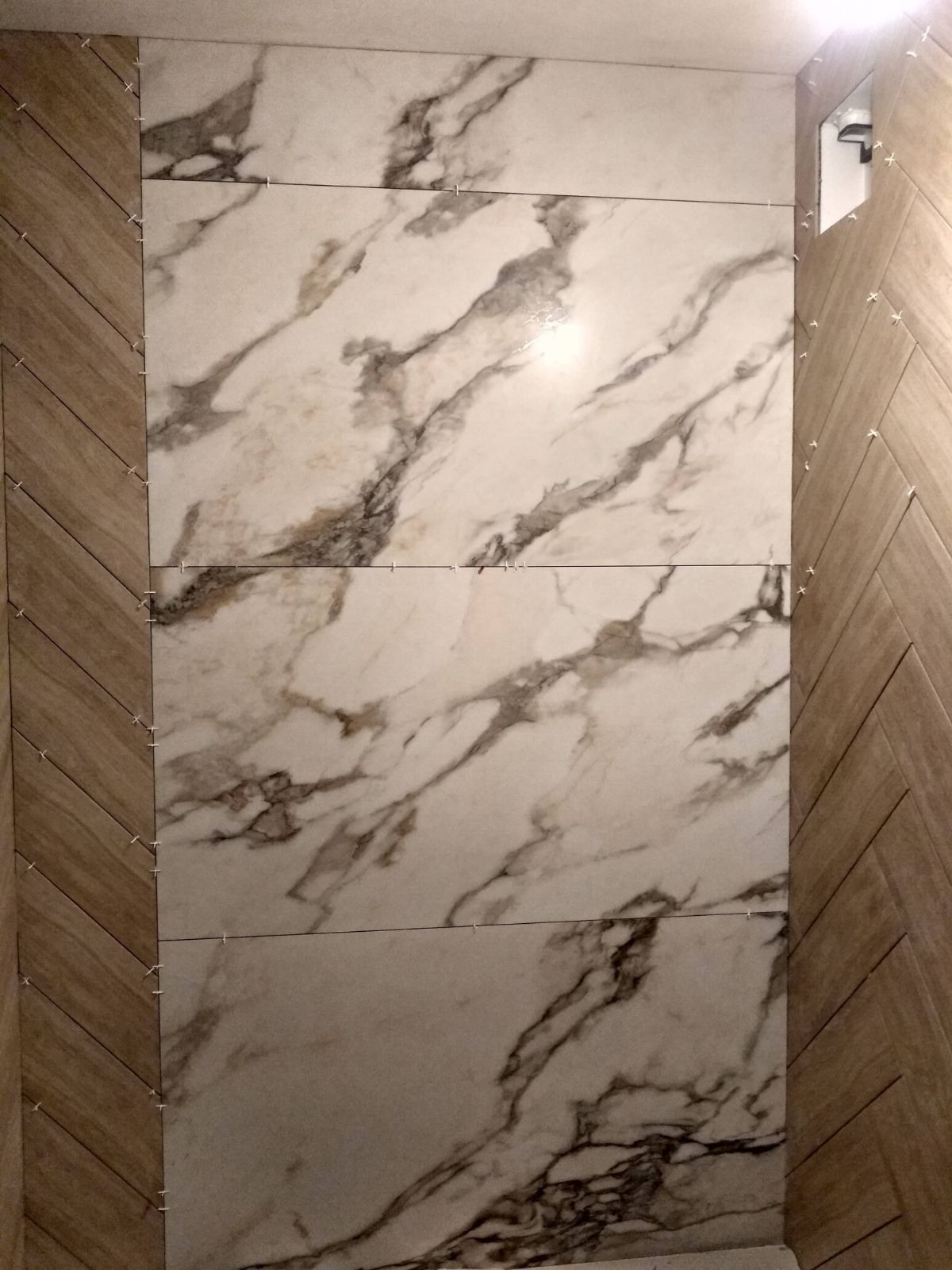 Ściana wyłożona płytkami imitującymi marmur, z drewnopodobnymi panelami ułożonymi w jodełkę po bokach. Widoczne krzyżyki dystansowe.