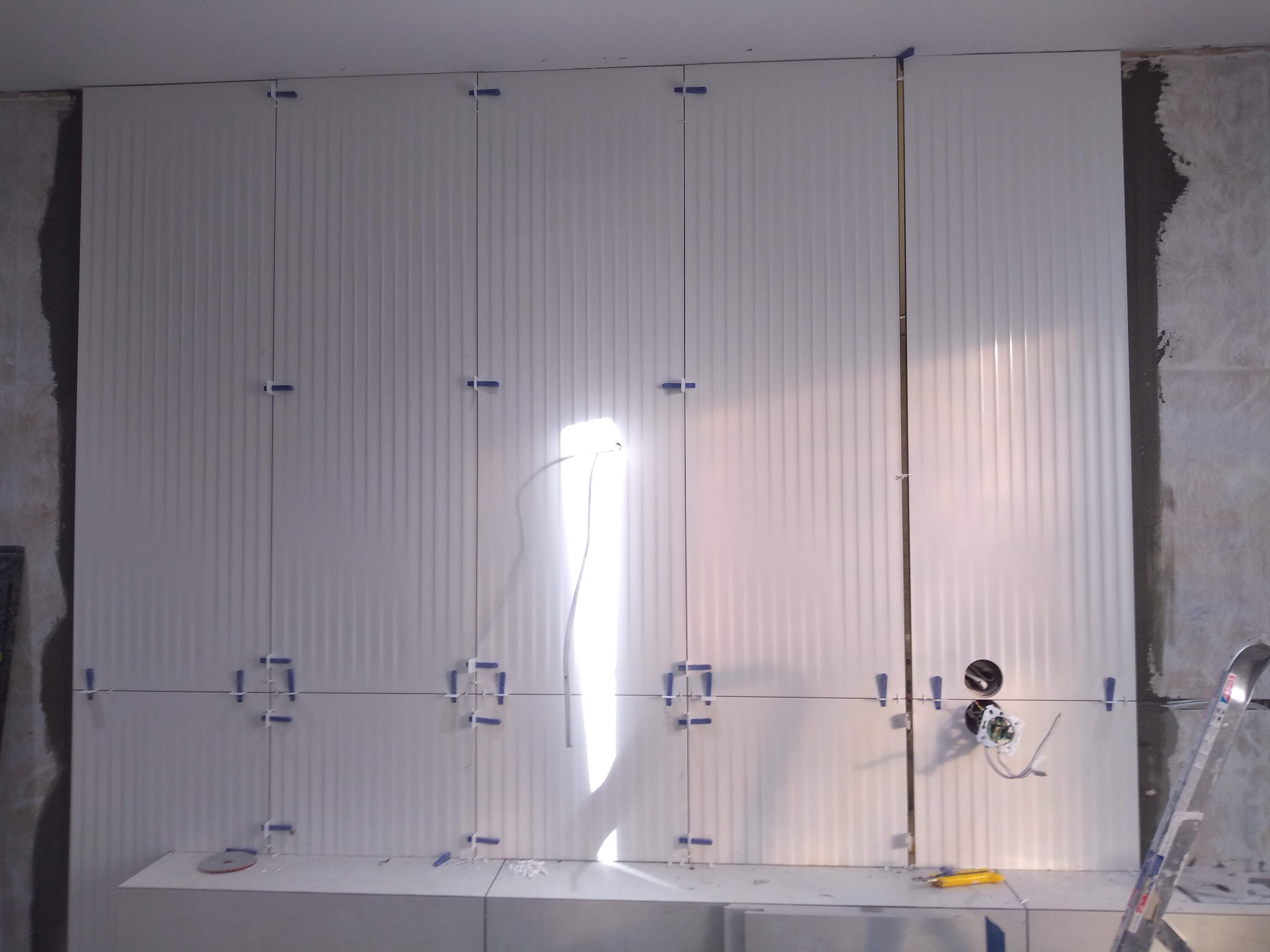 Białe, pionowo żłobkowane płytki ścienne, częściowo ułożone, z niebieskimi krzyżykami dystansowymi. Widoczne otwory na gniazdka i fragment ściany przed wykończeniem.