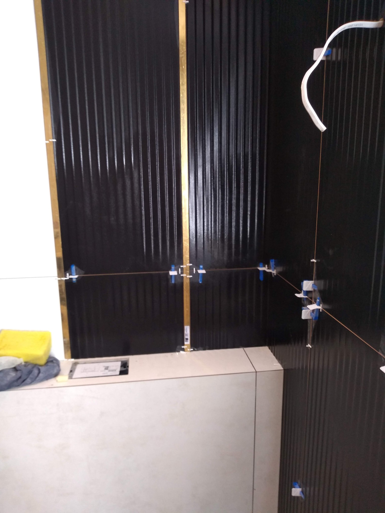 Montaż czarnych, pionowych paneli ściennych z użyciem poziomicy laserowej i klipsów. Widoczny fragment białej umywalki i akcesoria glazurnicze.