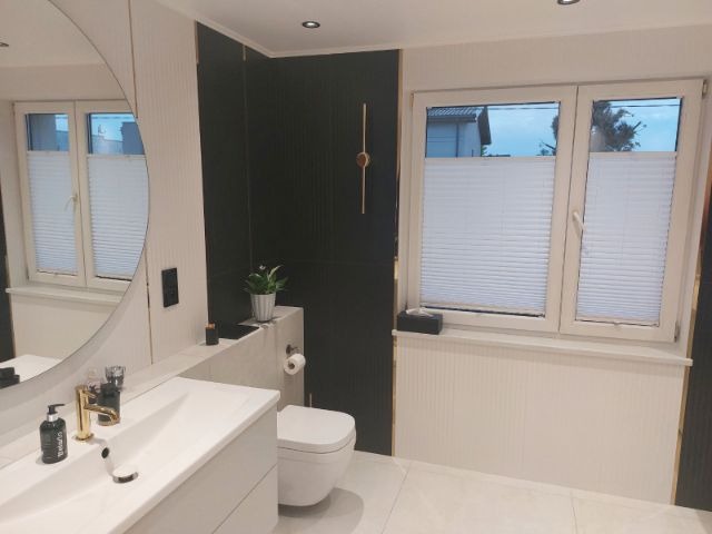 Nowoczesna łazienka z czarnymi panelami, białymi płytkami i owalnym lustrem. Złota armatura i rolety plisowane w oknach dodają elegancji.