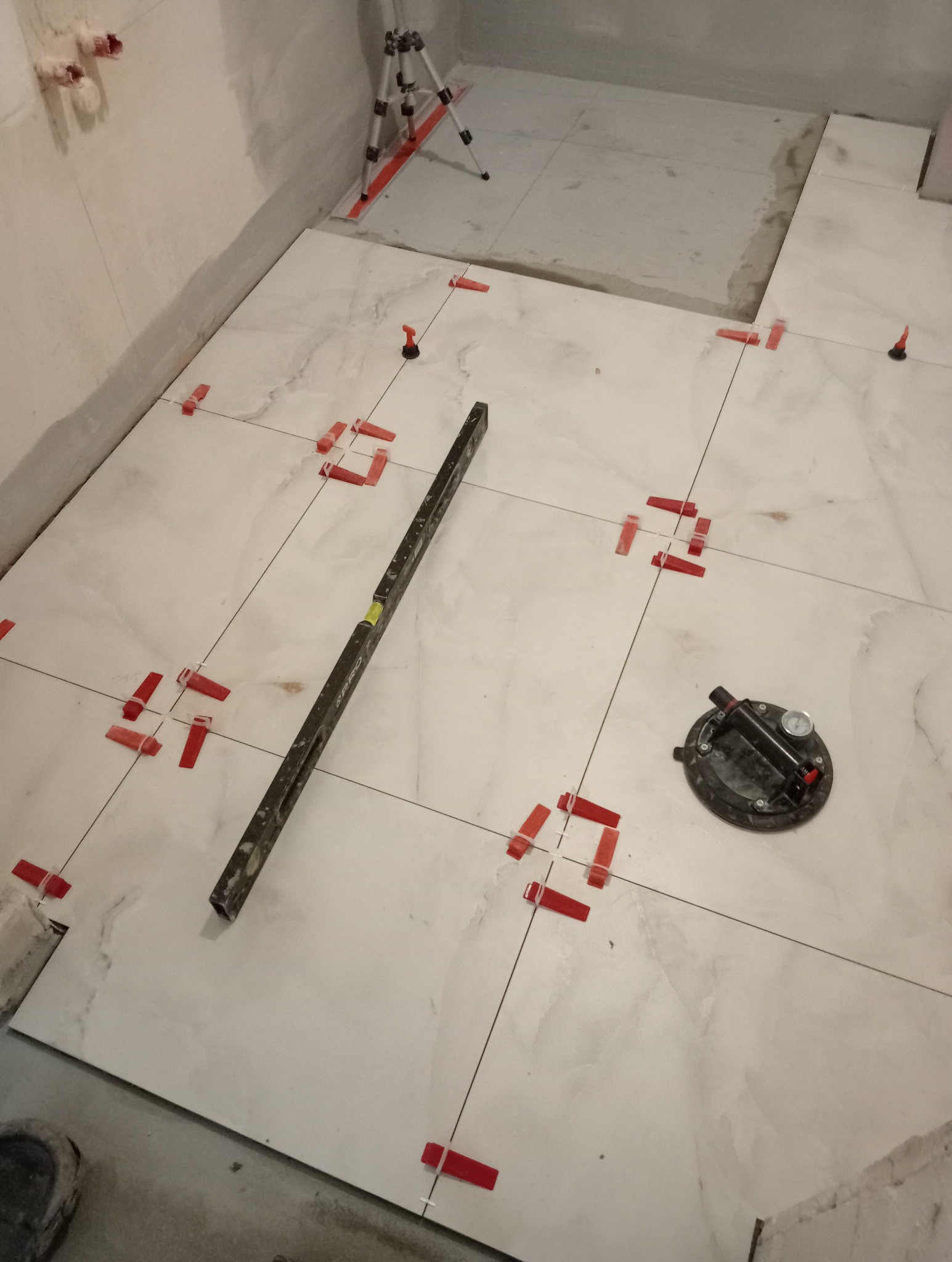 Białe płytki podłogowe w trakcie układania, z czerwonymi klipsami poziomującymi, poziomica i przyssawka do płytek. Widoczne rury i fragment niezakończonej podłogi.
