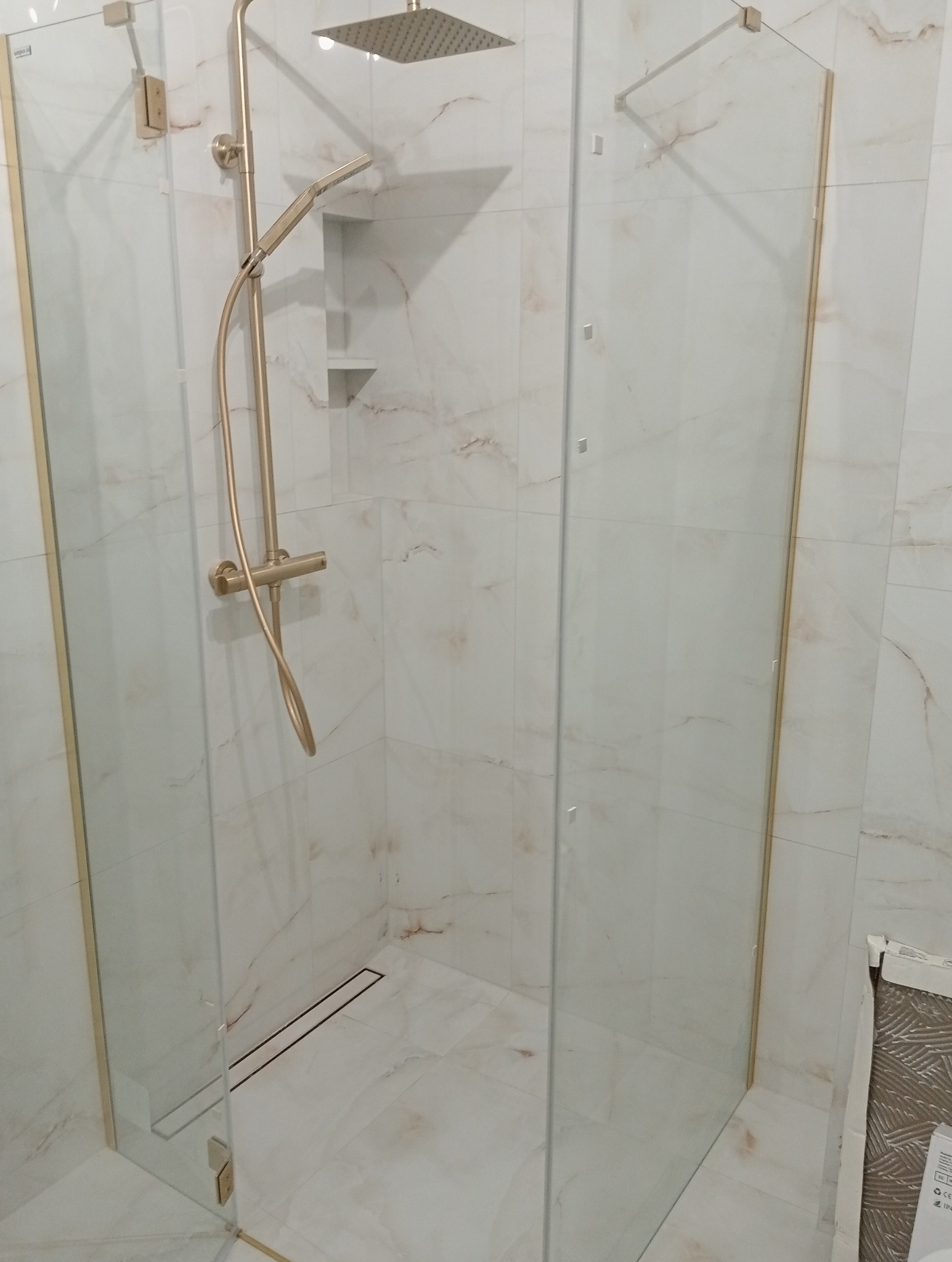Kabina prysznicowa walk-in z armaturą w kolorze złota, wykończona płytkami imitującymi marmur. Widoczny odpływ liniowy i półki w ścianie.