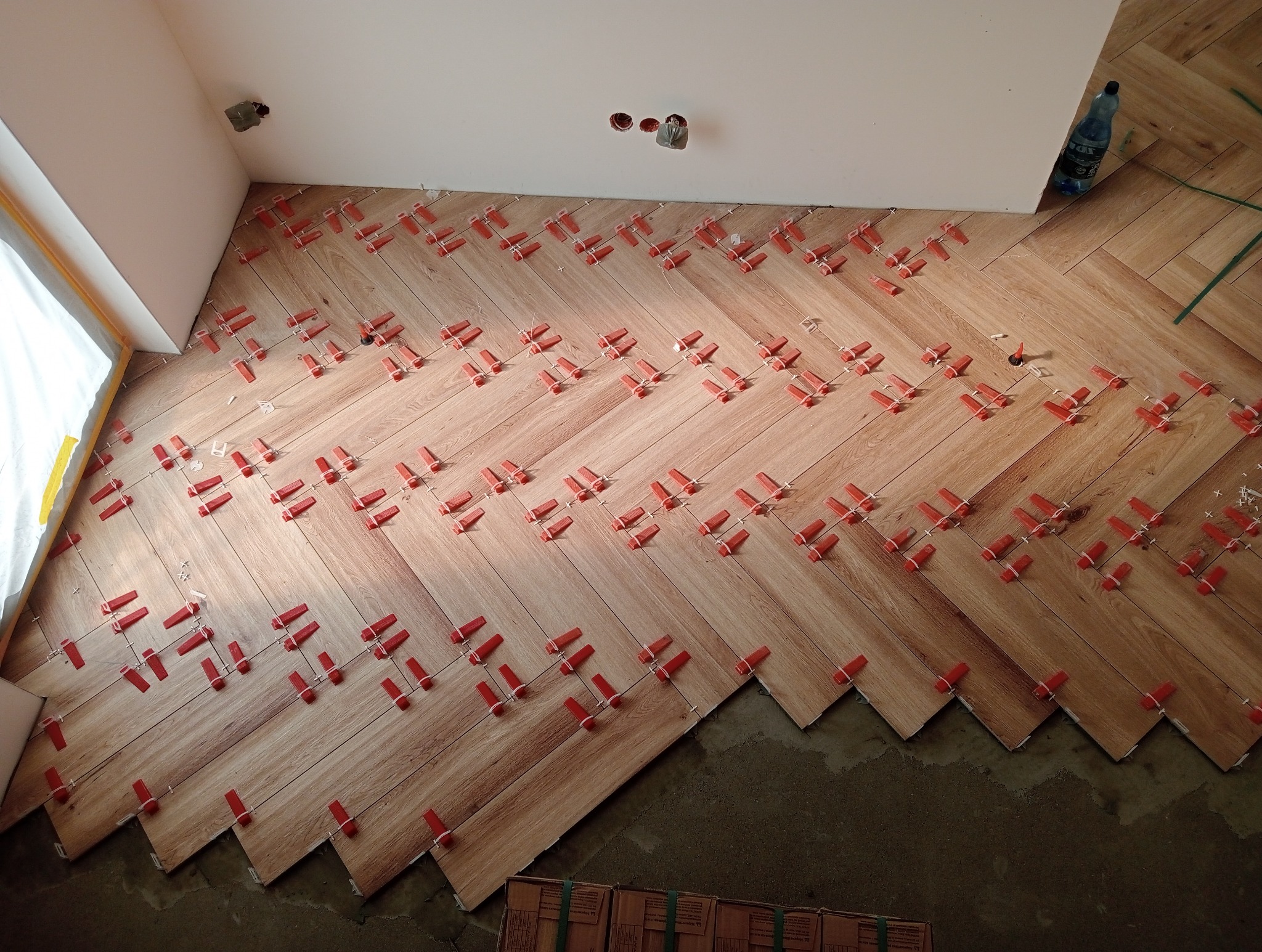 Ułożone płytki podłogowe w jodełkę, z czerwonymi klipsami poziomującymi. Widoczne niedociągnięcia przy ścianie i resztki kleju. Butelka wody w rogu.