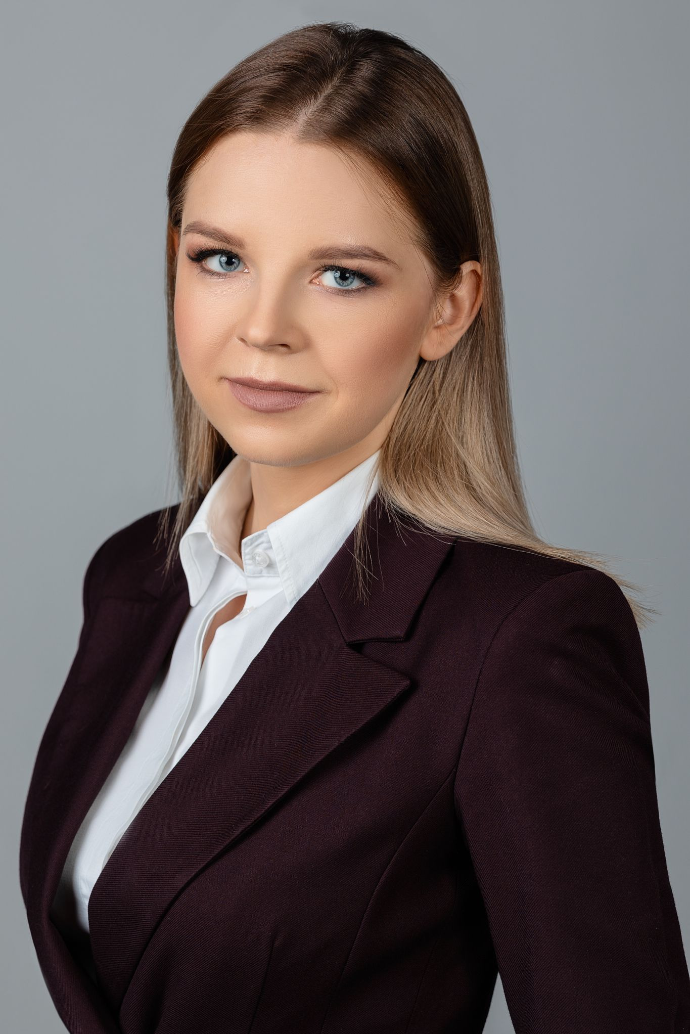 Portret kobiety z jasnymi włosami i niebieskimi oczami, ubrana w białą koszulę i bordową marynarkę na szarym tle.