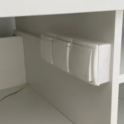 Białe, potrójne gniazdko elektryczne z symbolem dzwonka, zamontowane wewnątrz białej szafki, widoczny fragment żółto-zielonego przewodu.