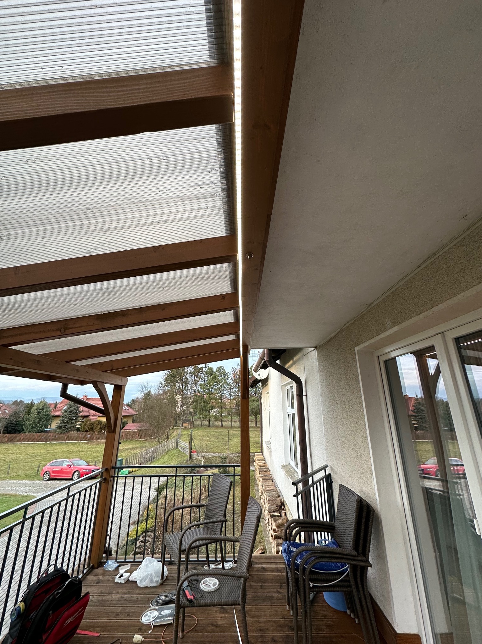 Zadaszony taras z drewnianą konstrukcją i poliwęglanowym dachem, oświetlony taśmą LED, widoczne krzesła, narzędzia i elementy instalacyjne na podłodze.