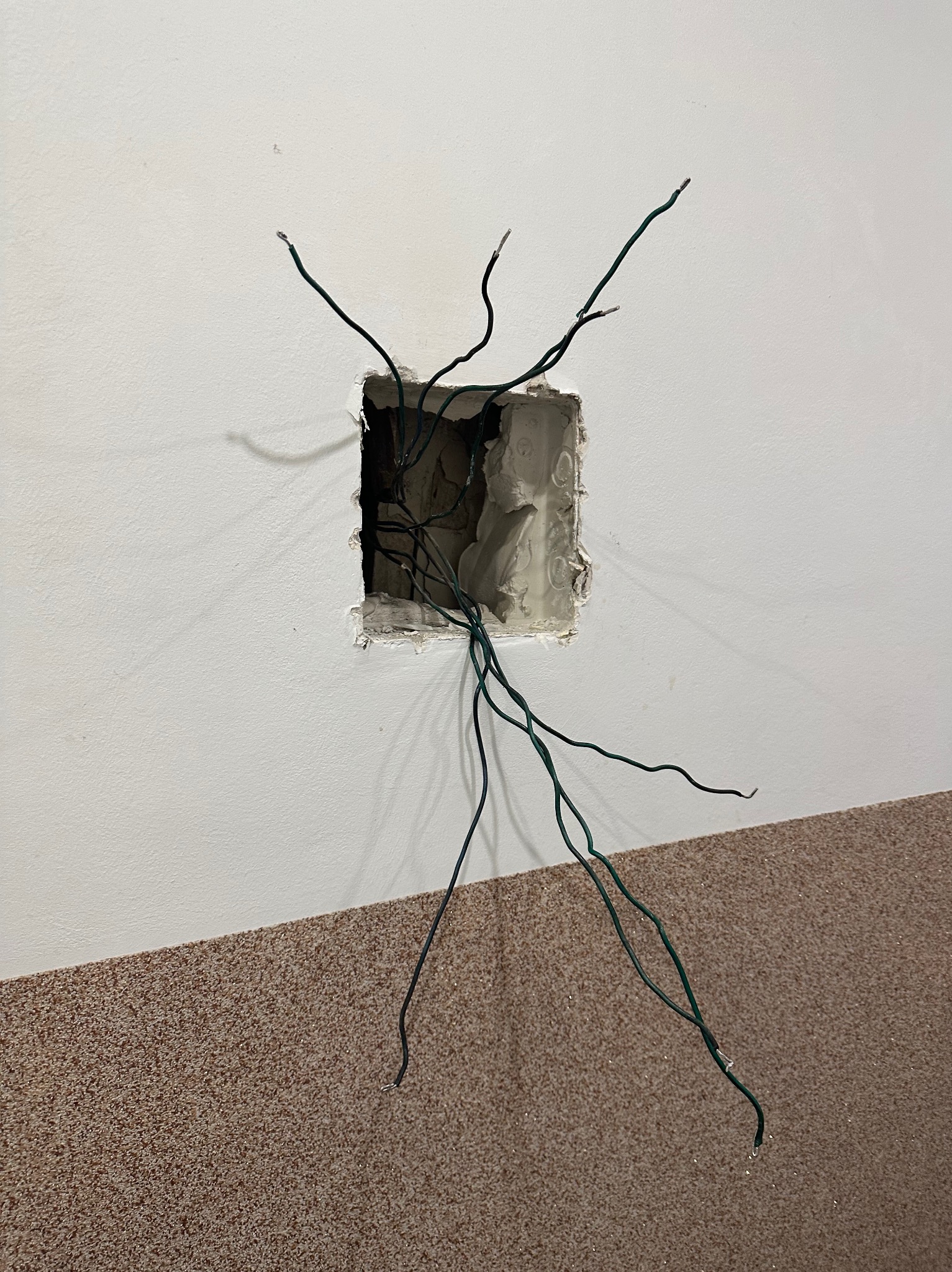 Otwór w ścianie z wystającymi zielonymi przewodami elektrycznymi, widoczne ślady gipsu wokół otworu, fragment ściany pomalowany na biało, dolna część ściany wykończona wykładziną dywanową...