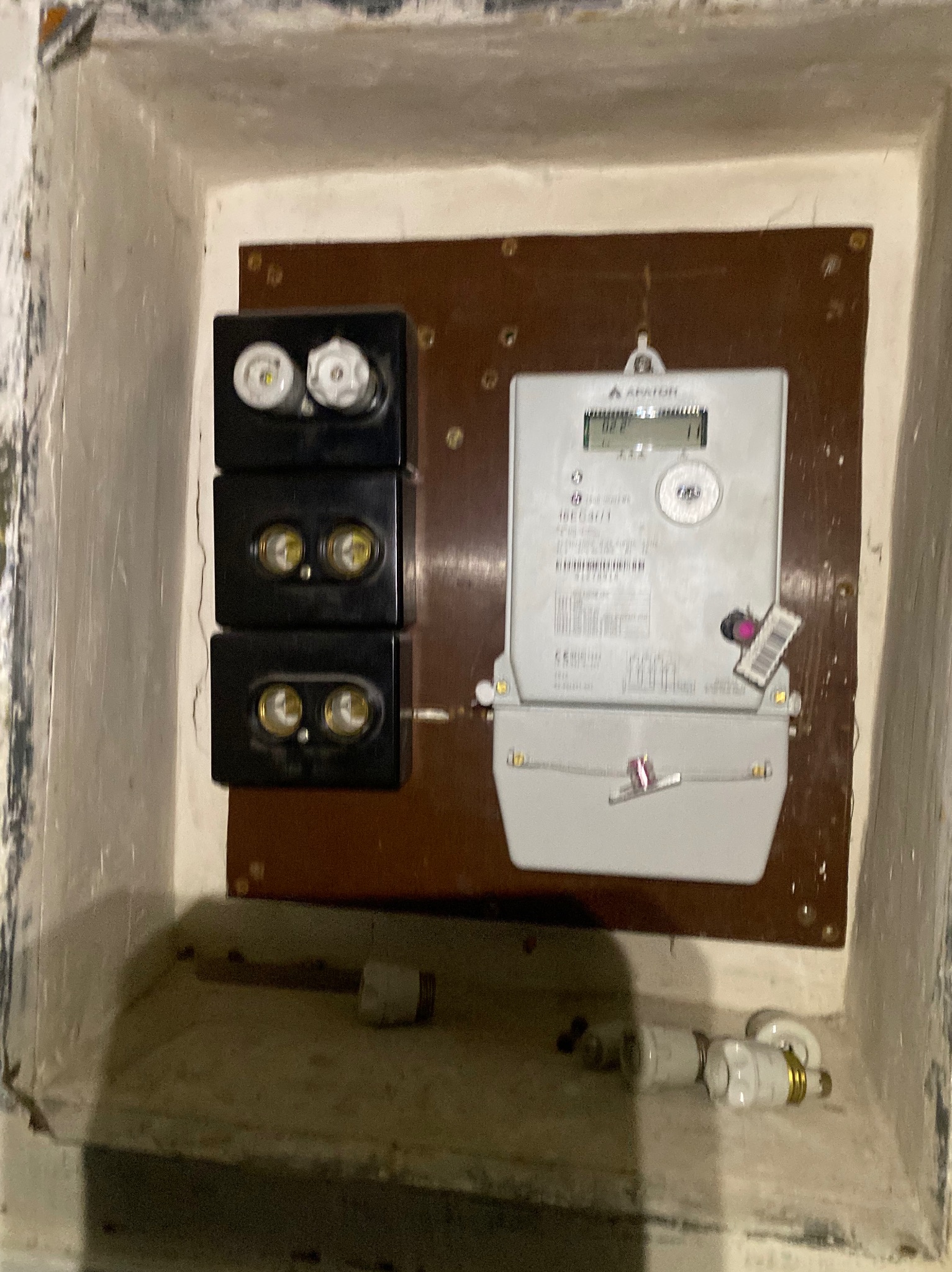 Zamontowany licznik elektryczny na brązowej płycie z trzema czarnymi bezpiecznikami w białej wnęce, widoczne dodatkowe bezpieczniki na dolnej półce.