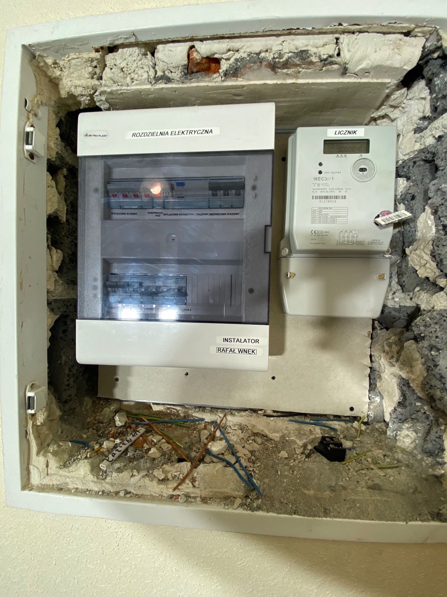 Rozdzielnia elektryczna i licznik zamontowane we wnęce w surowym stanie, widoczne przewody, gruz i oznaczenie instalatora Rafała Wnęka.