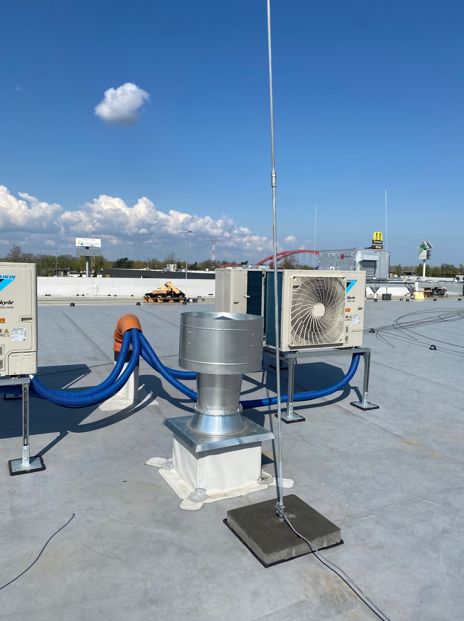 Instalacja klimatyzacji Daikin SkyAir z niebieskimi rurami na płaskim dachu, widoczny komin wentylacyjny i maszt odgromowy z betonową podstawą.
