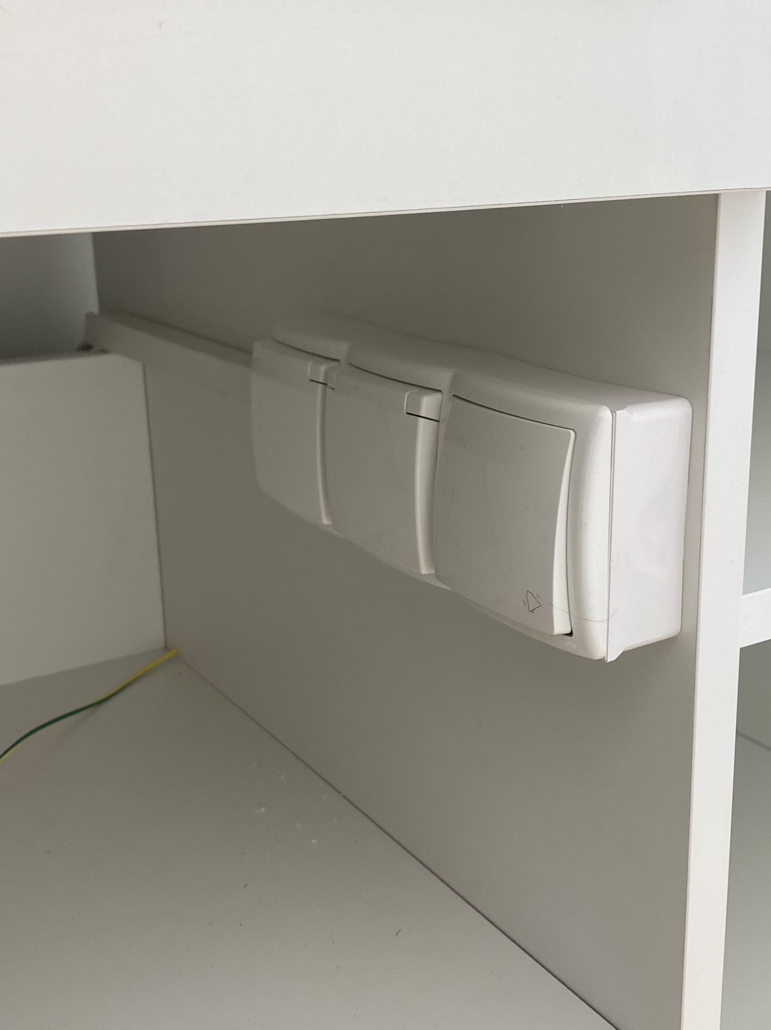 Białe, potrójne gniazdko elektryczne z symbolem dzwonka, zamontowane wewnątrz białej szafki, widoczny fragment żółto-zielonego przewodu.