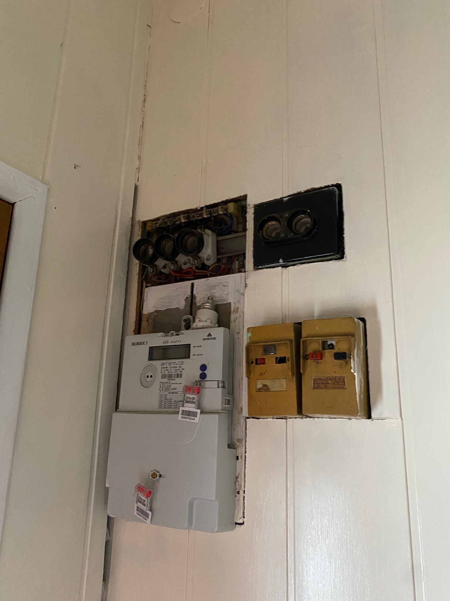 Szczegółowe ujęcie elektrycznego licznika Norax 3 i starych bezpieczników w drewnianej obudowie na białej ścianie z boazerią.