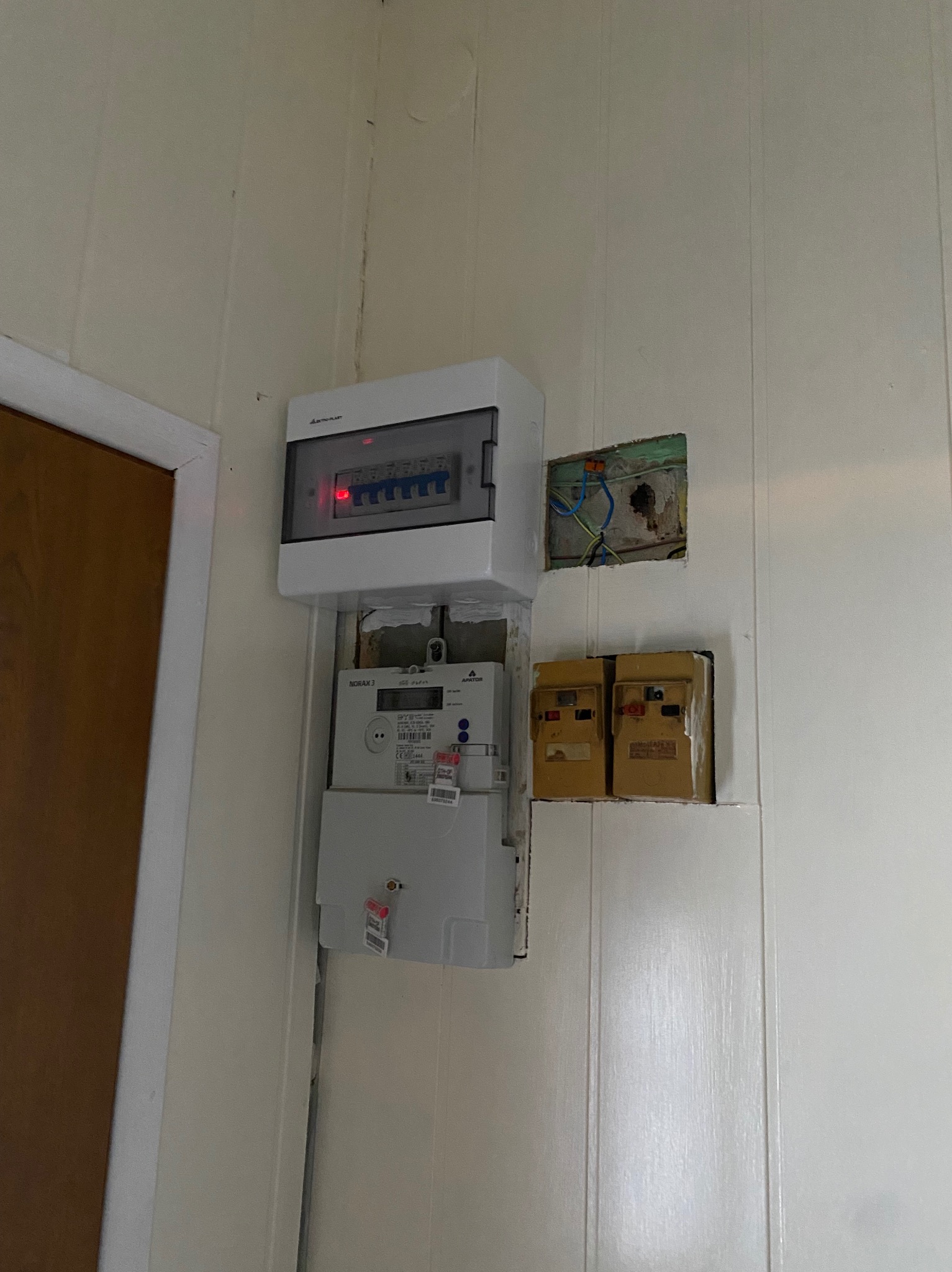 Biała skrzynka z bezpiecznikami nad licznikiem prądu na ścianie z boazerią, obok widoczne puszki instalacyjne z przewodami.