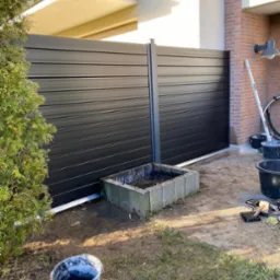 Nowoczesna, pozioma brama wjazdowa z czarnych paneli kompozytowych, zamontowana w Pile, widoczna z perspektywy trawnika z donicą z betonu i narzędziami montażowymi.