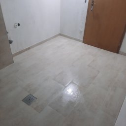 Zakład Usług Leśnych Robert Kowalski,Usługi budowlane - Beżowe, prostokątne płytki podłogowe w łazience z odpływem liniowym w rogu pomieszczenia, przy ścianie widoczne gniazdka elektryczne, a w tle drewniane drzwi.