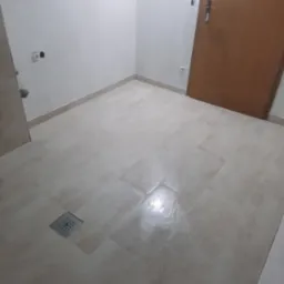 Beżowe, prostokątne płytki podłogowe w łazience z odpływem liniowym w rogu pomieszczenia, przy ścianie widoczne gniazdka elektryczne, a w tle drewniane drzwi.