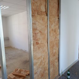 Zakład Usług Leśnych Robert Kowalski,Usługi budowlane - Montaż izolacji z wełny mineralnej w metalowym stelażu ściany działowej, widoczne fragmenty podłogi i sufitu z oświetleniem rastrowym, butelka wody przy ścianie.