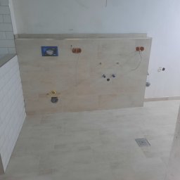 Zakład Usług Leśnych Robert Kowalski,Usługi budowlane - Ściana w trakcie prac wykończeniowych w łazience z widocznymi przyłączami hydraulicznymi, płytkami imitującymi kamień na ścianie i podłodze, oraz fragmentem ściany wyłożonej białymi, prostokątnymi...