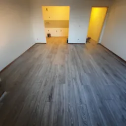 Ułożone szare panele podłogowe w pustym pokoju z widocznymi drzwiami do kuchni i innego pomieszczenia, ściany białe, widok z perspektywy podłogi.