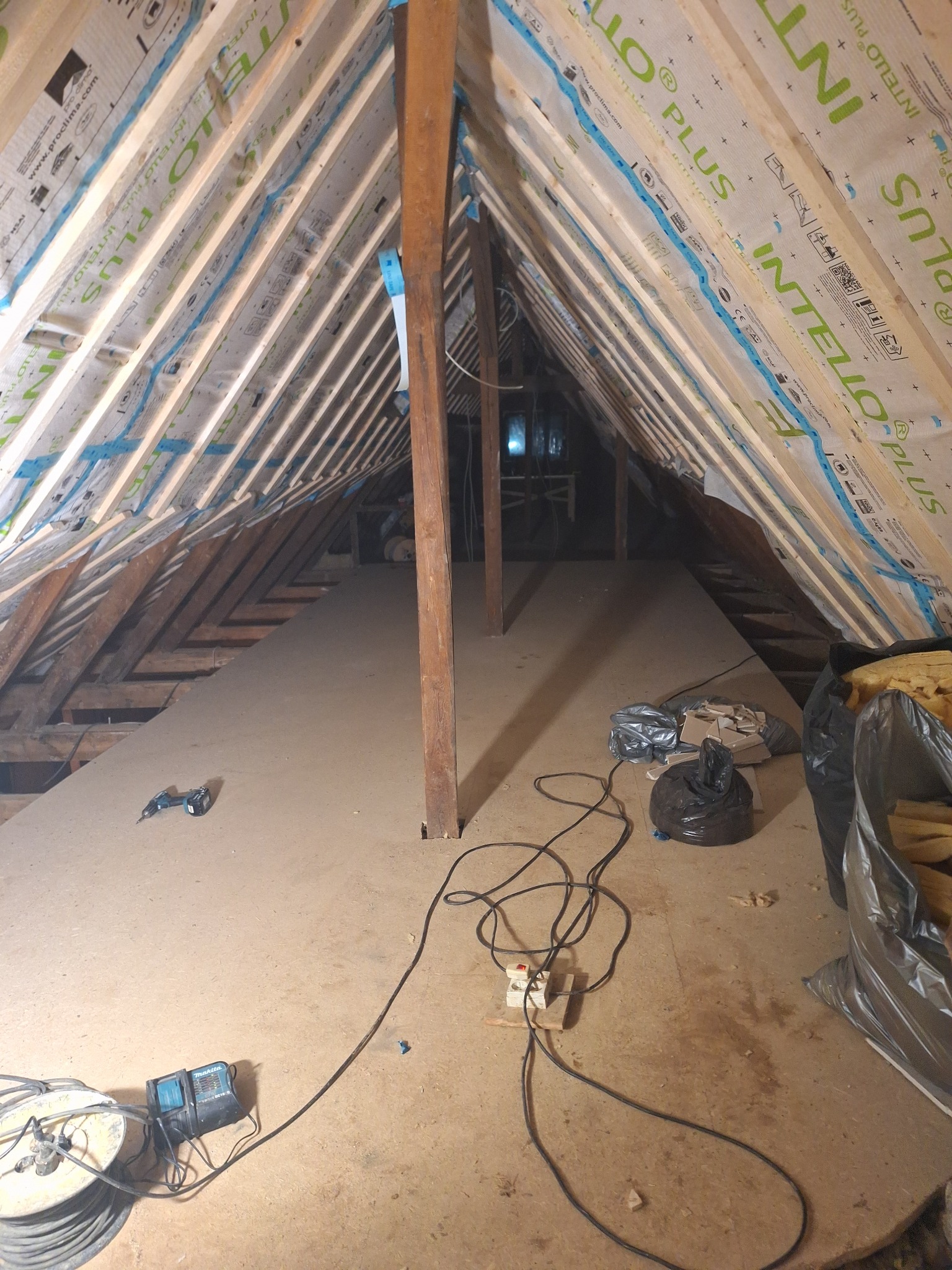 Poddasze w trakcie adaptacji: widoczne ocieplenie folią paroizolacyjną z nadrukiem 'INTELLO PLUS', drewniana konstrukcja dachu, płyty podłogowe, narzędzia (wiertarka akumulatorowa) i worki...