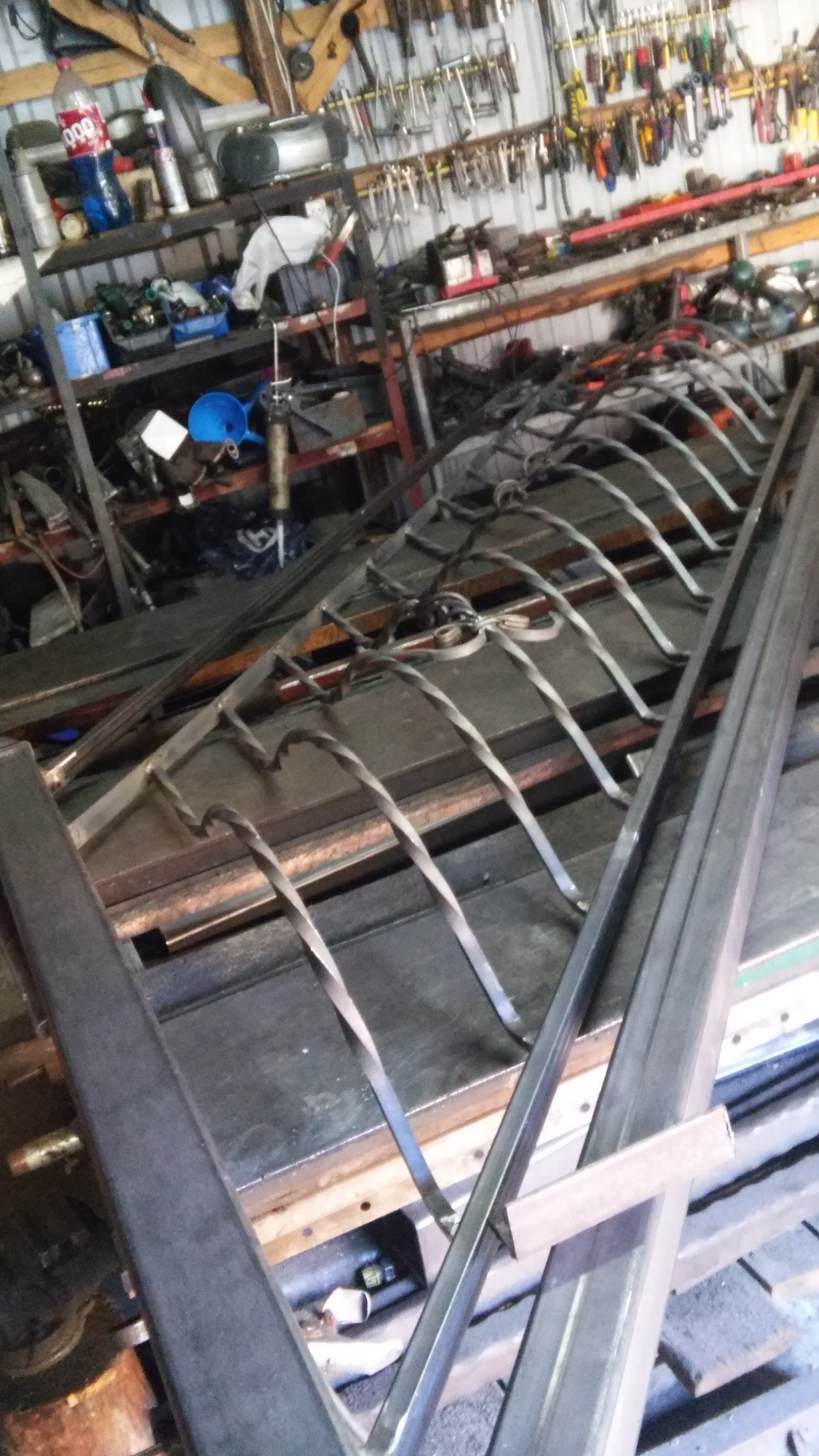 Fragment metalowej konstrukcji z ozdobnymi, skręconymi elementami, leżącej na warsztatowym stole, w tle widoczne narzędzia i wyposażenie warsztatu.