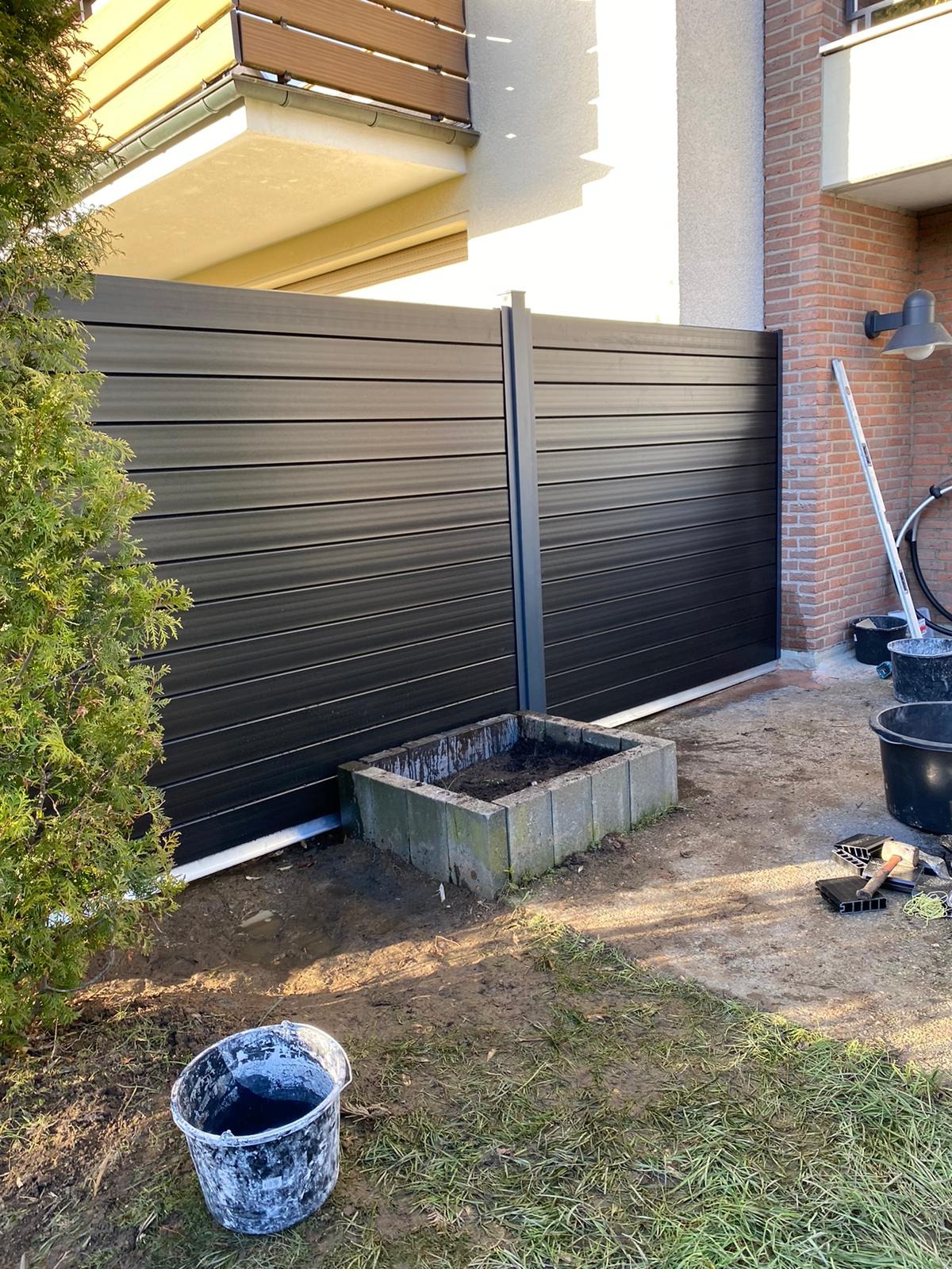 Nowoczesna, pozioma brama wjazdowa z czarnych paneli kompozytowych, zamontowana w Pile, widoczna z perspektywy trawnika z donicą z betonu i narzędziami montażowymi.