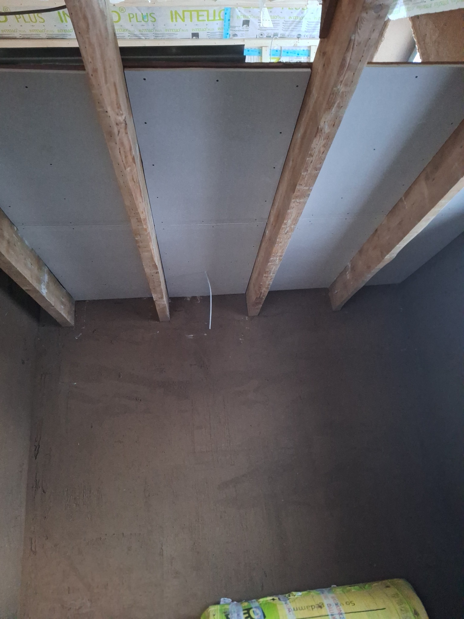 Surowy pokój w trakcie remontu: drewniane belki stropowe, płyty gipsowe na suficie i ściany pokryte tynkiem glinianym. Widoczny kabel elektryczny zwisający ze stropu oraz fragment opakowania...