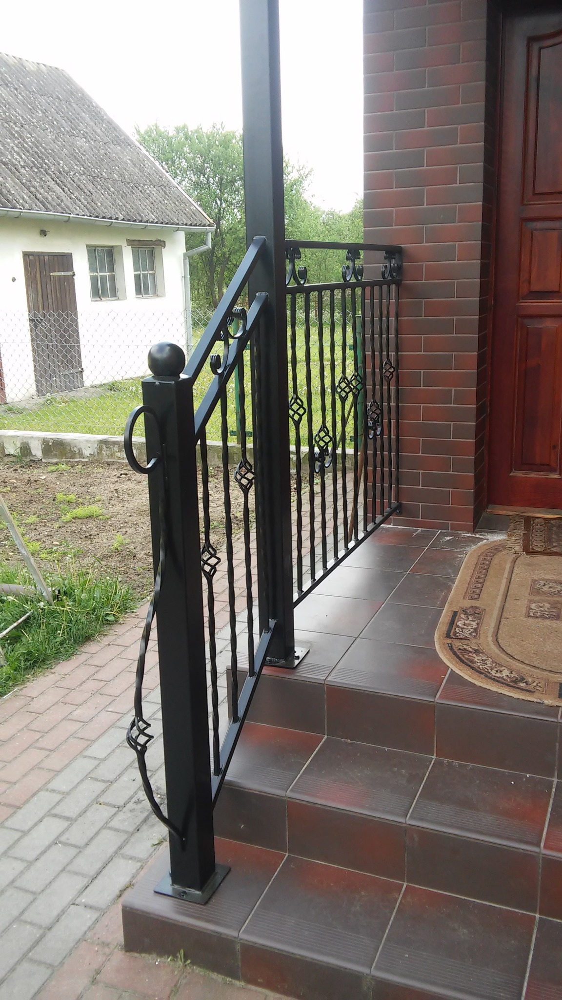 Czarne, metalowe schody zewnętrzne z ozdobnymi elementami, prowadzące do drzwi wejściowych z brązowej cegły.