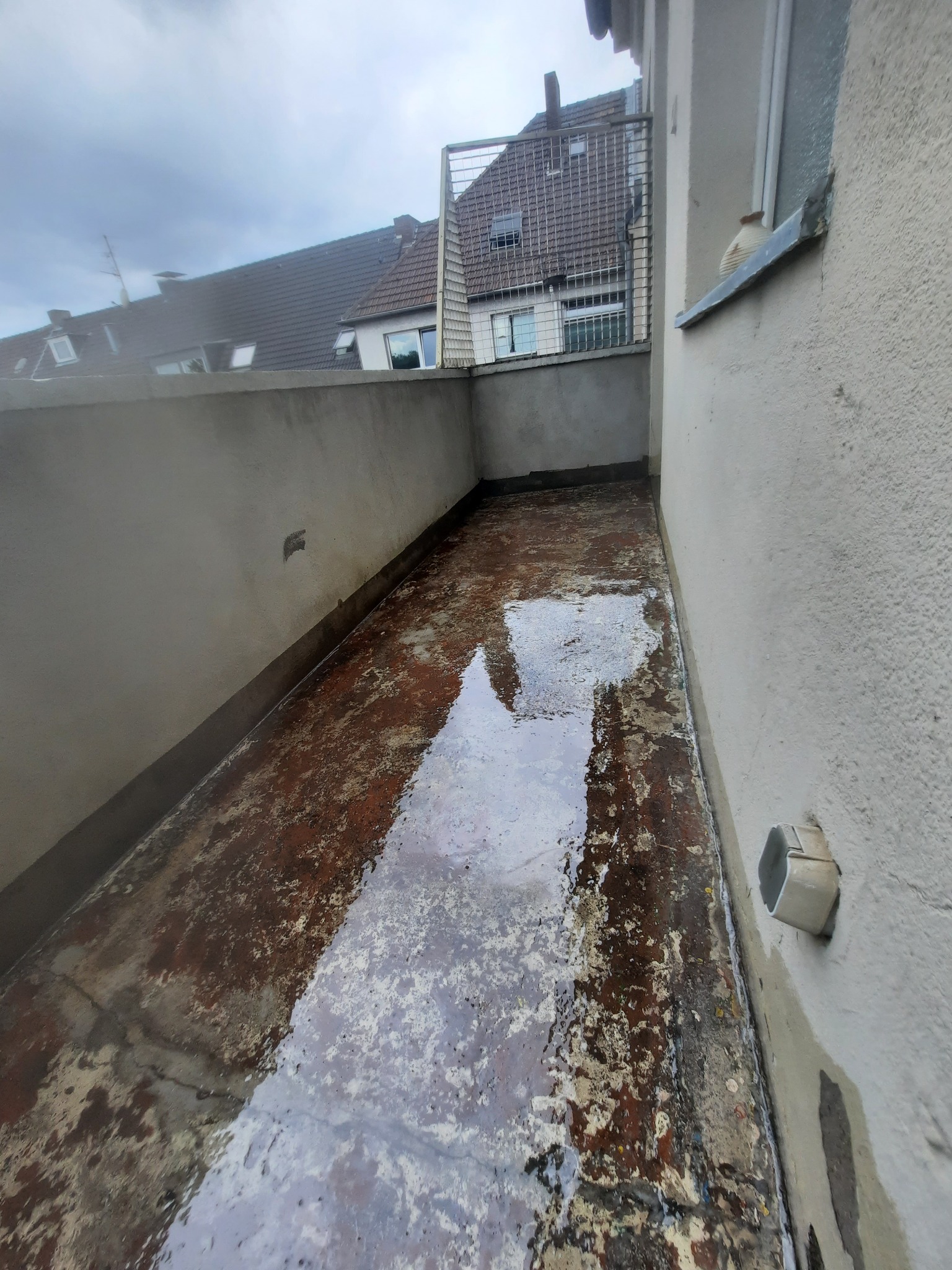 Zalany, brudny balkon z widocznymi śladami starej farby i wody, z szarymi ścianami i fragmentem budynku w tle.
