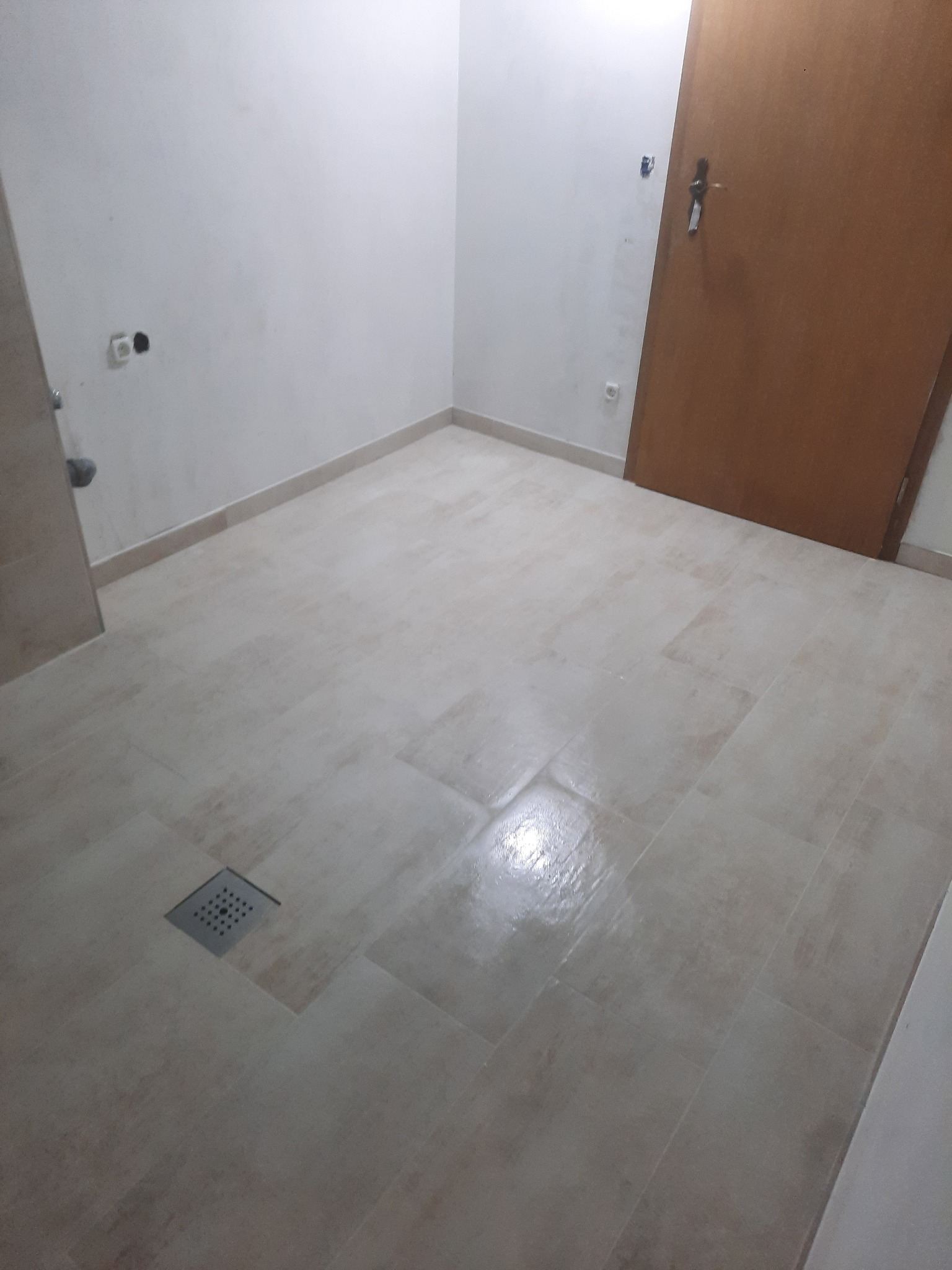 Beżowe, prostokątne płytki podłogowe w łazience z odpływem liniowym w rogu pomieszczenia, przy ścianie widoczne gniazdka elektryczne, a w tle drewniane drzwi.