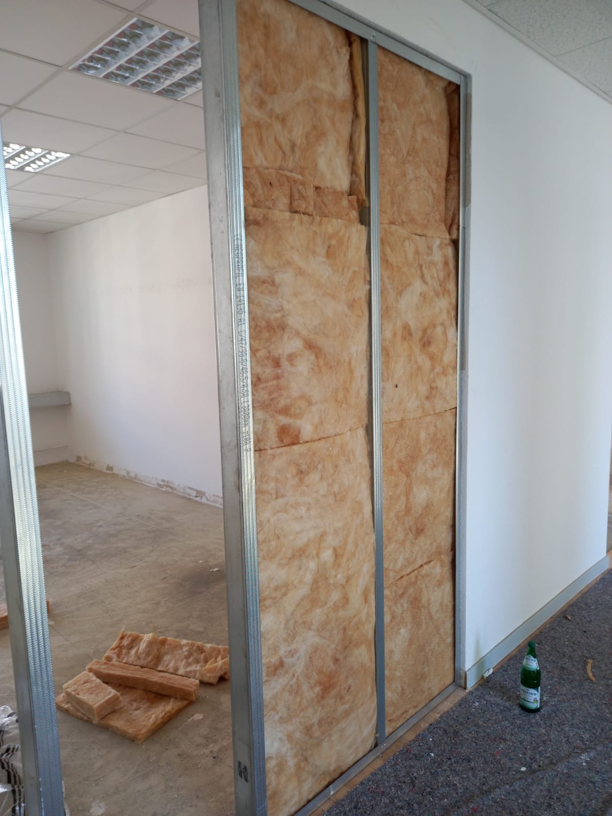 Montaż izolacji z wełny mineralnej w metalowym stelażu ściany działowej, widoczne fragmenty podłogi i sufitu z oświetleniem rastrowym, butelka wody przy ścianie.