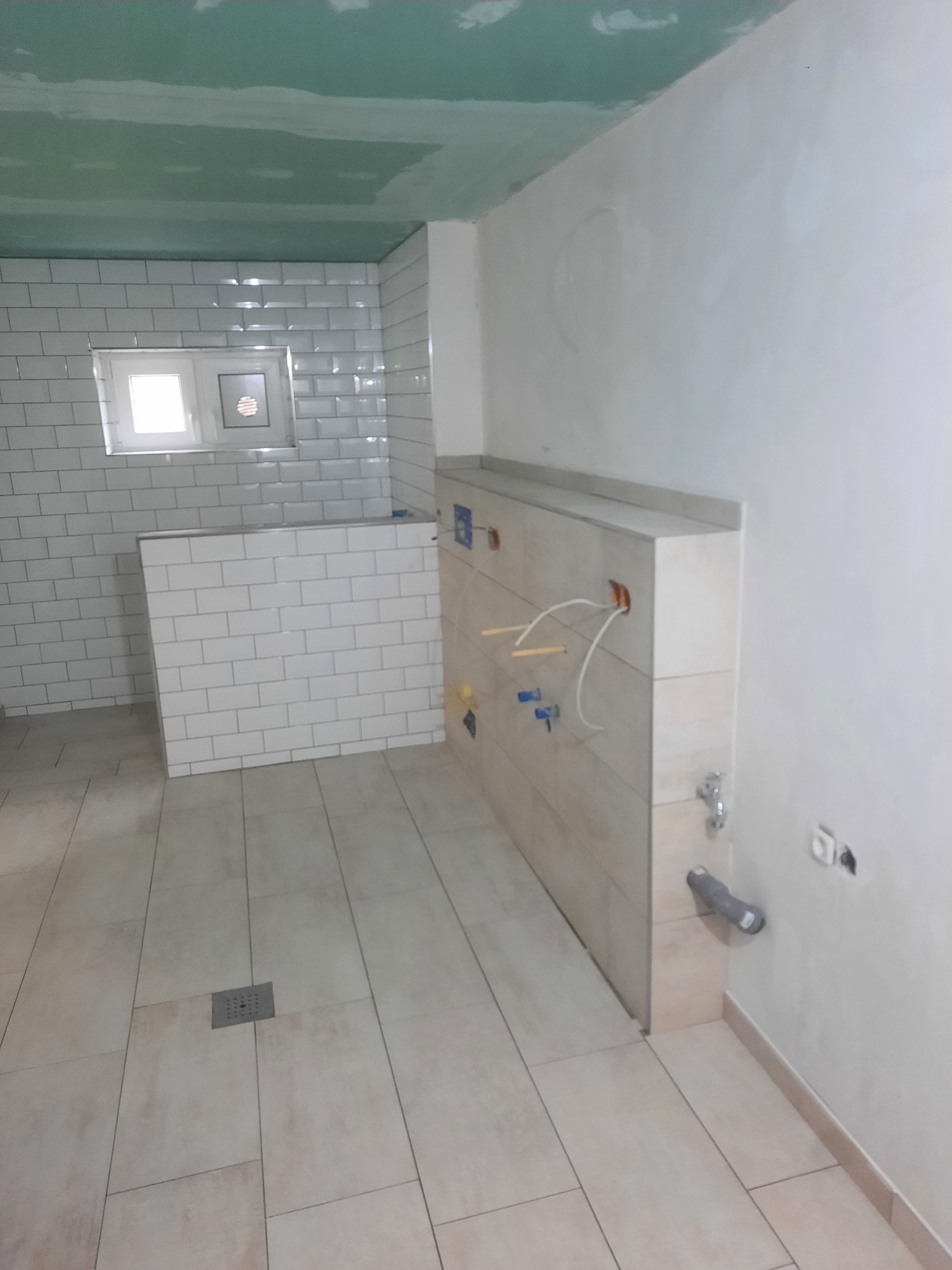Wnętrze łazienki w trakcie remontu: ułożone płytki podłogowe i ścienne typu metro, widoczne instalacje wodne i elektryczne, sufit w trakcie malowania.