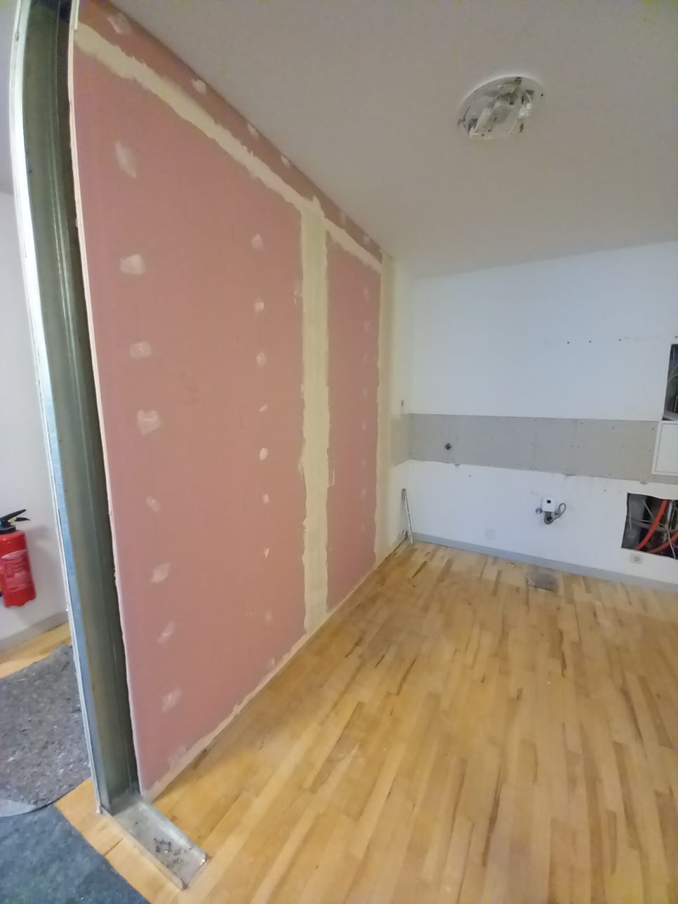 Wnętrze w trakcie prac wykończeniowych: ściana z płyt gipsowo-kartonowych z widocznymi łączeniami szpachlowanymi, drewniana podłoga, częściowo wykończona ściana z instalacją elektryczną, czerwona...