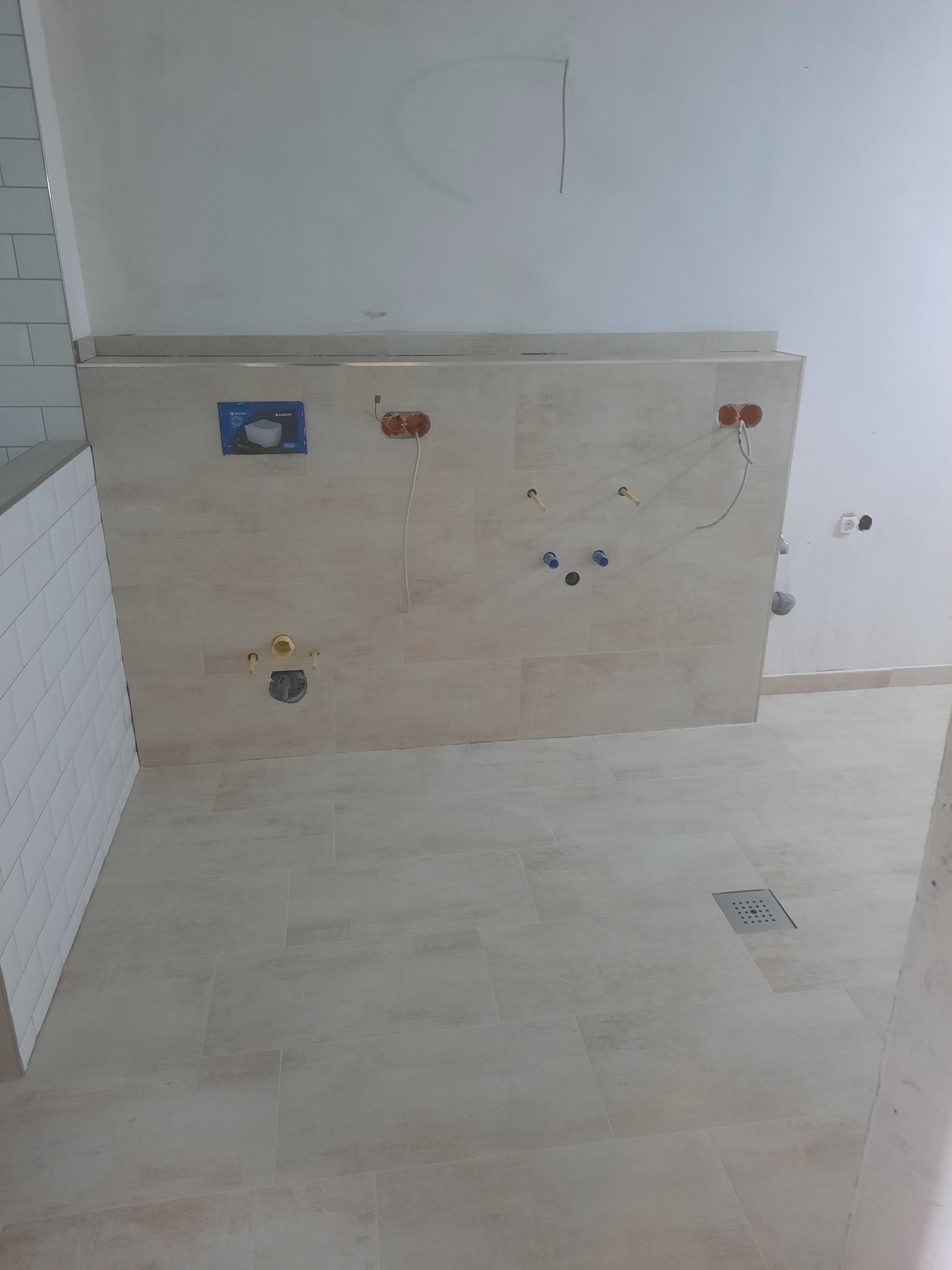 Ściana w trakcie prac wykończeniowych w łazience z widocznymi przyłączami hydraulicznymi, płytkami imitującymi kamień na ścianie i podłodze, oraz fragmentem ściany wyłożonej białymi, prostokątnymi...