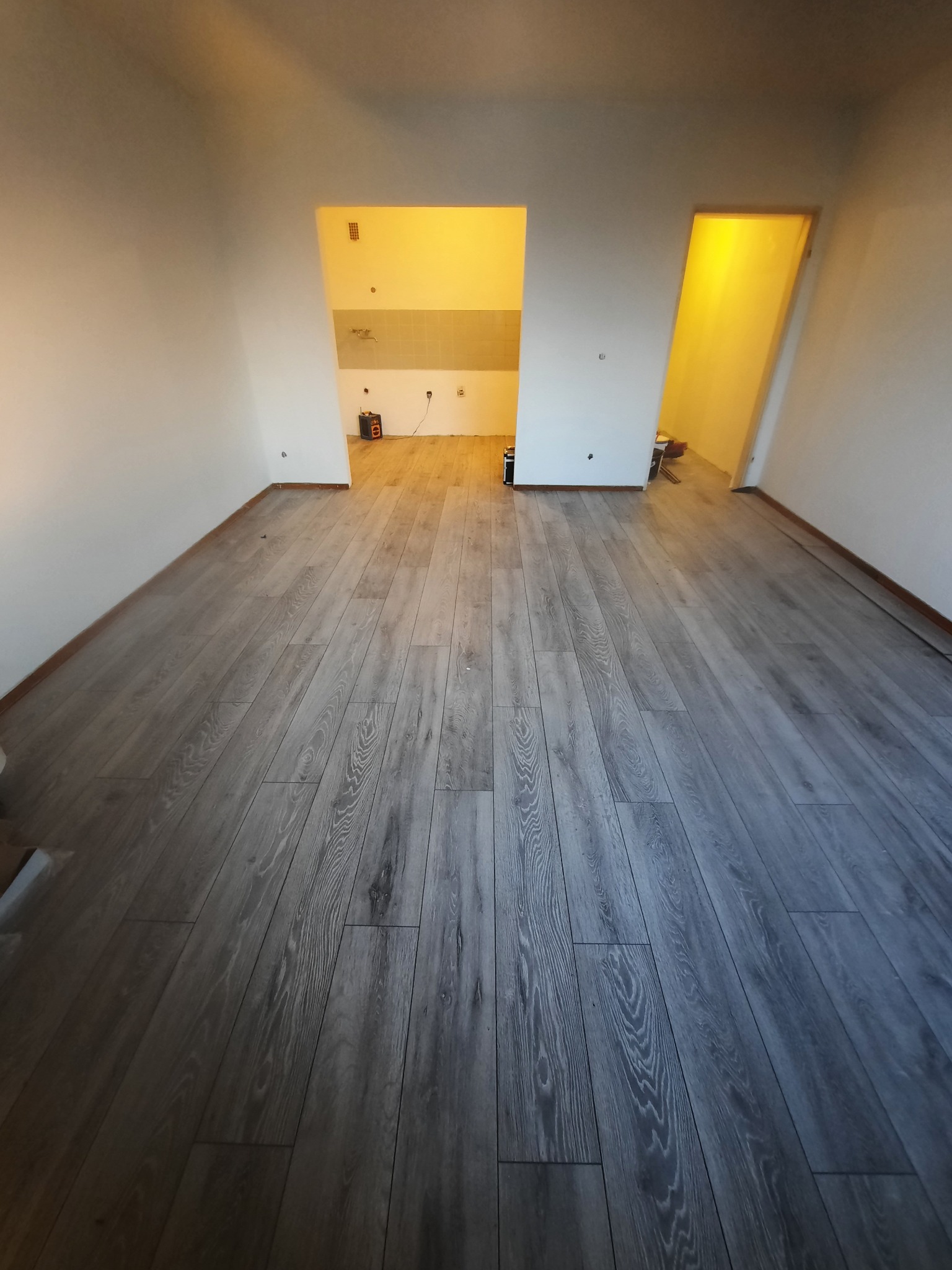 Ułożone szare panele podłogowe w pustym pokoju z widocznymi drzwiami do kuchni i innego pomieszczenia, ściany białe, widok z perspektywy podłogi.