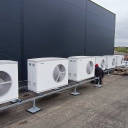 TomekekoDomek - Rząd białych jednostek zewnętrznych pomp ciepła marki Stiebel Eltron, zamontowanych na metalowych konstrukcjach na dachu budynku przemysłowego, instalator w czerwonej bluzie dokonuje podłączeń