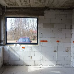 TomekekoDomek - Surowy stan wnętrza budynku w Białymstoku z widocznymi instalacjami elektrycznymi w ścianach z betonu komórkowego i rurami wentylacyjnymi na podłodze, okno z widokiem na zewnątrz.