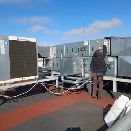 TomekekoDomek - Instalator w rękawicach montuje system wentylacyjny VTS na dachu budynku, widoczny również agregat klimatyzacyjny Climaveneta i fragment instalacji rurowej.