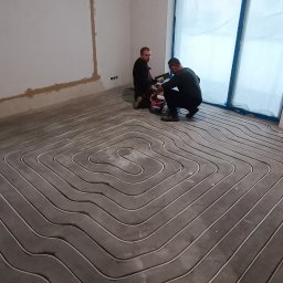TomekekoDomek - Dwóch instalatorów w czarnych ubraniach montuje rury ogrzewania podłogowego w pomieszczeniu z surowymi ścianami i oknem zabezpieczonym folią.