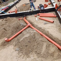 TomekekoDomek - Układanie pomarańczowych rur kanalizacyjnych w piasku na fundamencie budynku, widoczne narzędzia i sprzęt budowlany, prace ziemne w toku.