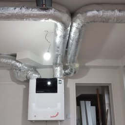 TomekekoDomek - Centrala wentylacyjna Zehnder ComfoAir Q z widocznymi kanałami wentylacyjnymi w izolacji aluminiowej, zamontowana w nowym budynku z białymi ścianami i oknem.