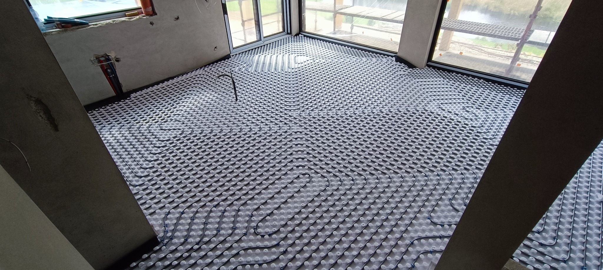 Instalacja ogrzewania podłogowego w surowym stanie, widoczne rury grzewcze ułożone w pętle na izolacji, okna balkonowe z widokiem na zewnątrz, fragment ściany z instalacją elektryczną.