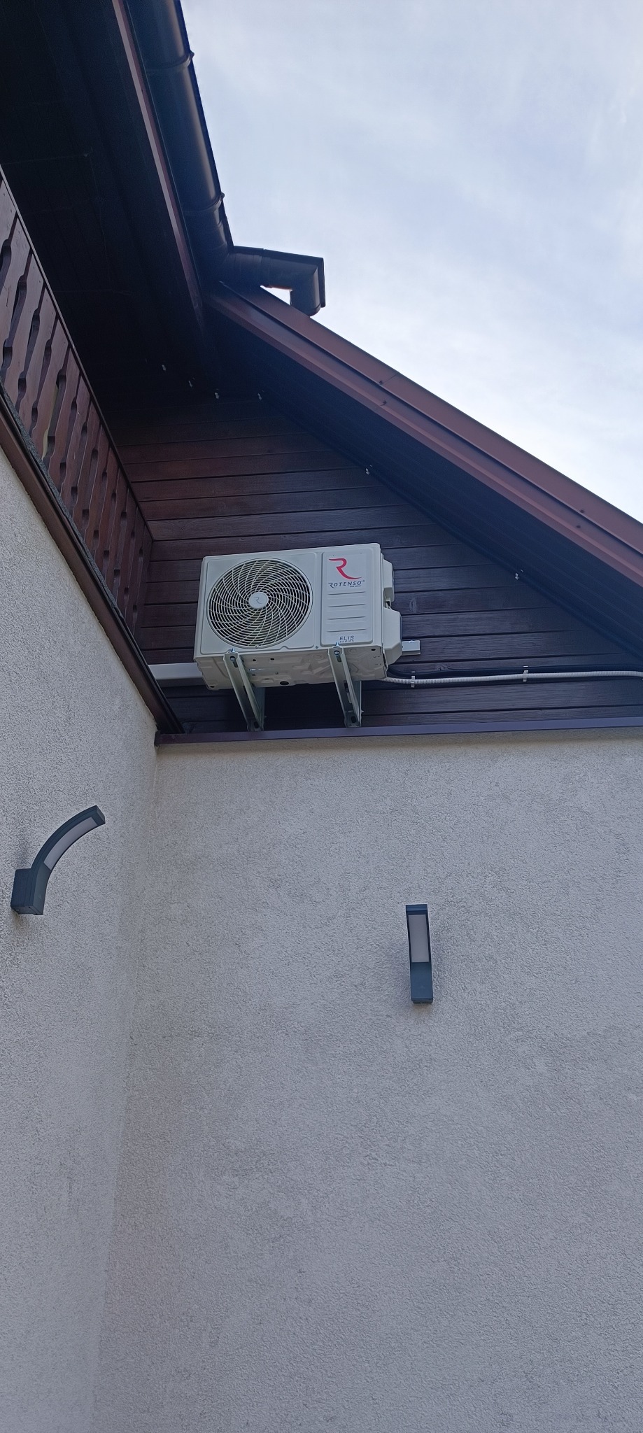 Zewnętrzna jednostka klimatyzacji marki Rotenso Elis Series zamontowana na wspornikach na elewacji domu z drewnianą podbitką dachową i dwoma kinkietami.