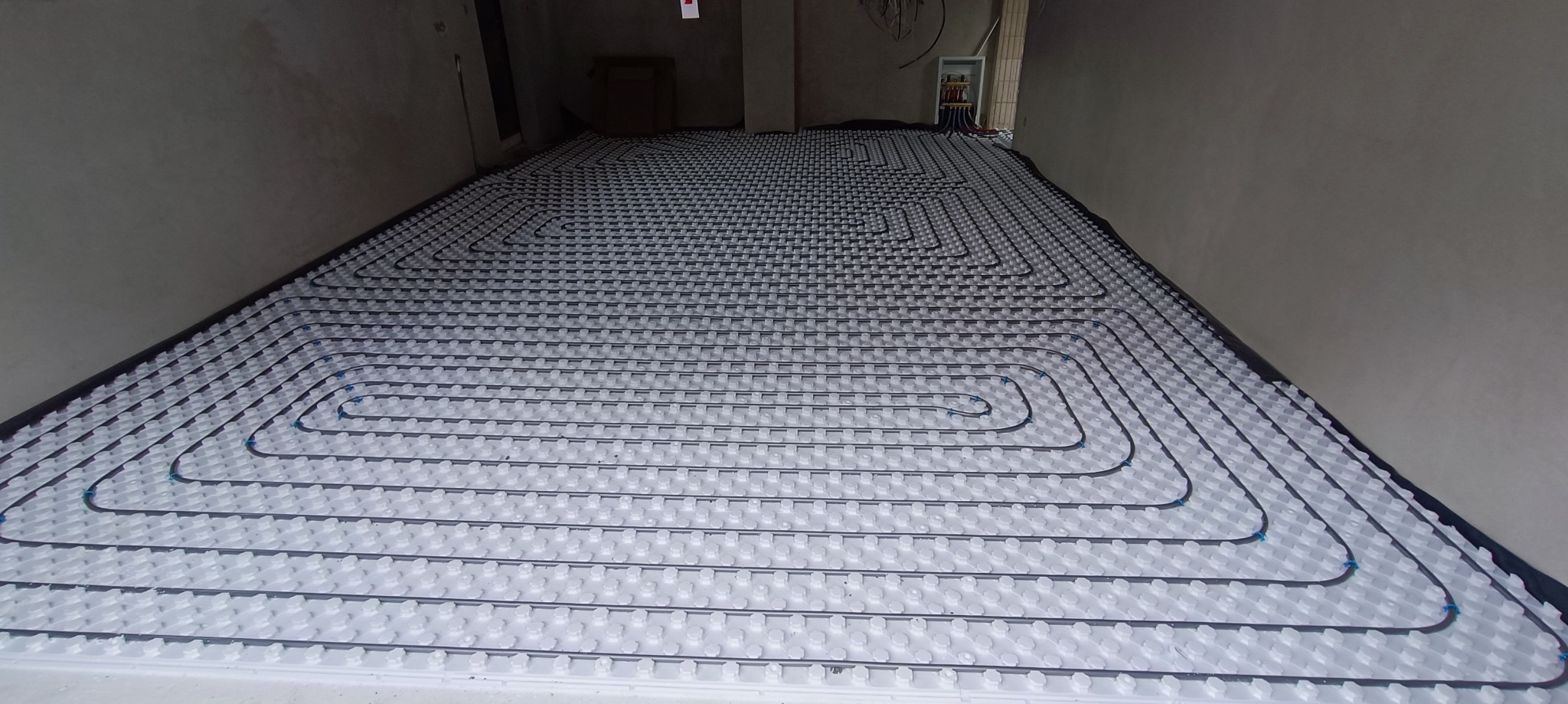 Rozłożona instalacja ogrzewania podłogowego z rurkami na białych płytach styropianowych w surowym wnętrzu, widok z góry, instalacja w trakcie montażu.