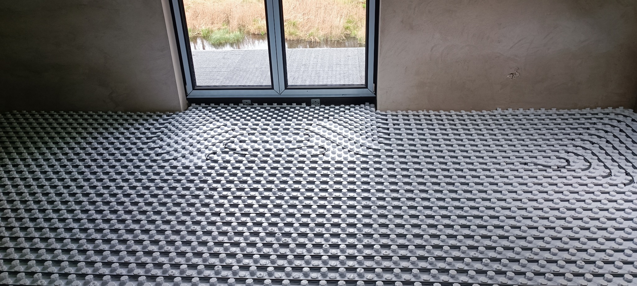 Instalacja ogrzewania podłogowego w nowym budynku, widoczne rury w systemie mocowań, okno z widokiem na zewnątrz, surowy tynk na ścianach.