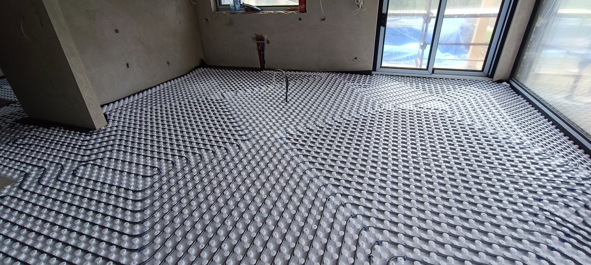 Rozłożone rury ogrzewania podłogowego na białej macie izolacyjnej w surowym pomieszczeniu z dużym oknem balkonowym.