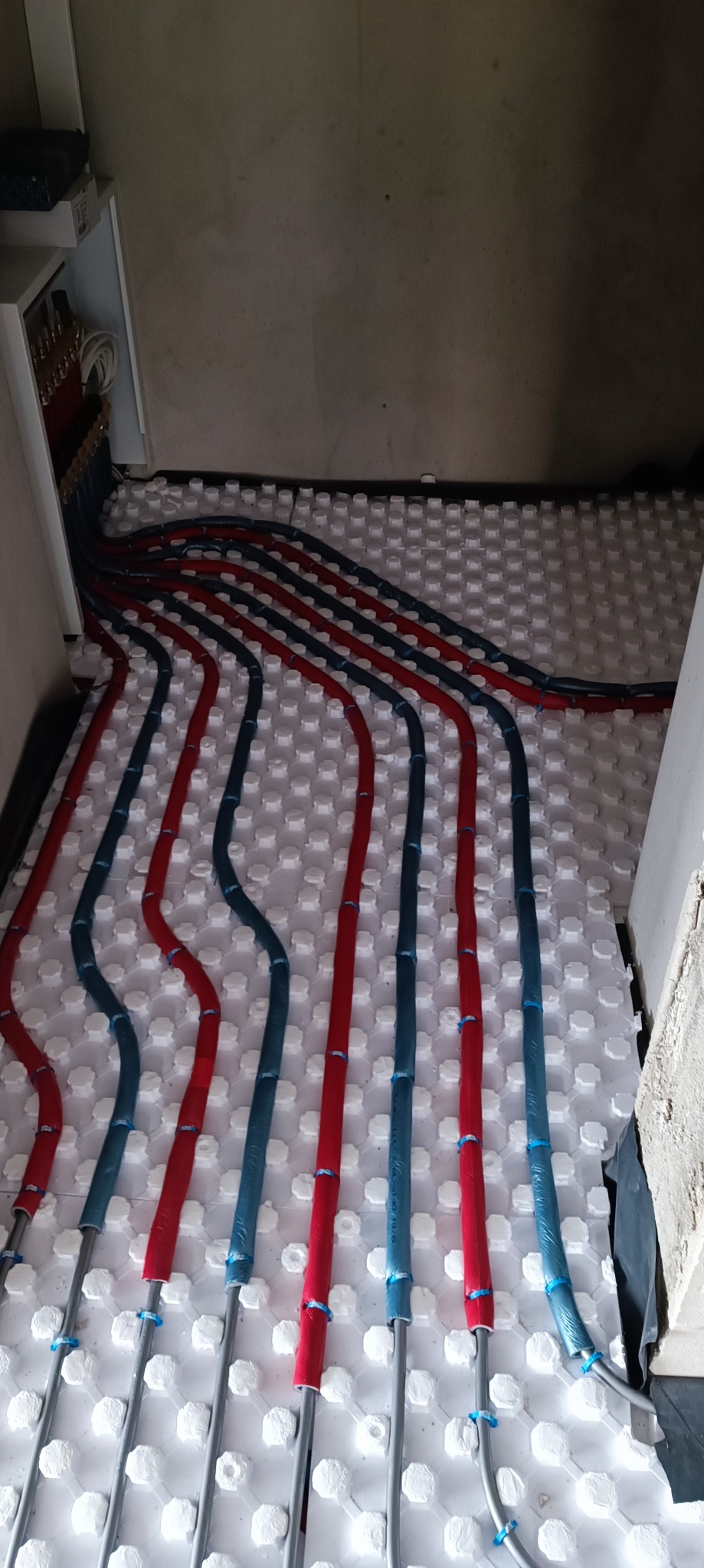 Instalacja ogrzewania podłogowego: rury w kolorach czerwonym i niebieskim ułożone na białych panelach izolacyjnych z widoczną rozdzielnią w tle.