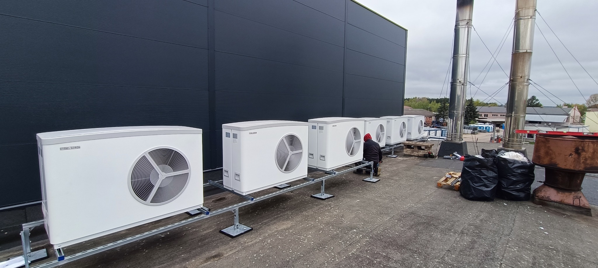 Rząd białych jednostek zewnętrznych pomp ciepła marki Stiebel Eltron, zamontowanych na metalowych konstrukcjach na dachu budynku przemysłowego, instalator w czerwonej bluzie dokonuje podłączeń