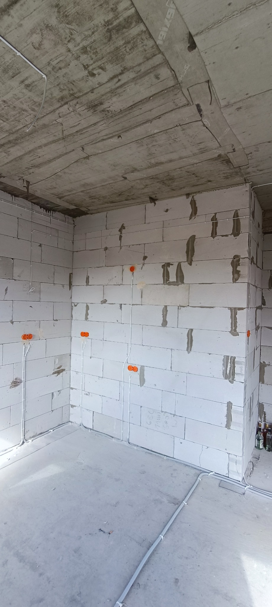 Surowy stan wnętrza z widoczną instalacją elektryczną: białe przewody prowadzone po ścianach z bloczków betonowych i podłodze, pomarańczowe puszki instalacyjne, surowy betonowy strop.